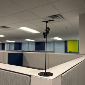 May include: A black, slender, vertical stand featuring a circular base, a thin central rod, and an adjustable mounting mechanism at the top. The device is designed for holding small items and is positioned upright on a flat surface.