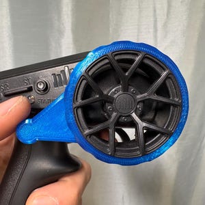 Può includere: Un dispositivo di controllo remoto nero e blu. Il dispositivo ha un'impugnatura nera e un componente blu a forma di ruota. La ruota ha un design nero a più razze. Il dispositivo è tenuto in mano.