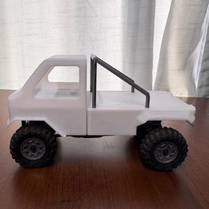 以下が含まれることがあります： 黒い車輪とタイヤを備えた、白い3Dプリントの小型ピックアップトラックのモデル。トラックにはロールバーとフラットベッドがあります。モデルは茶色の表面に置かれています。
