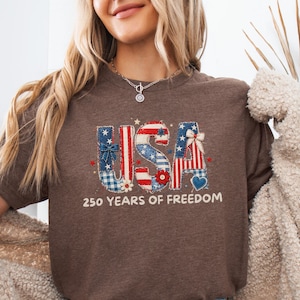 Puede incluir: Una camiseta marrón con la palabra "USA" en un diseño patriótico con patrones rojos, blancos y azules. El texto "250 YEARS OF FREEDOM" está impreso debajo. La camiseta se combina con pantalones cortos de mezclilla.