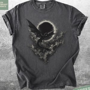 Puede incluir: Una camiseta gris carbón con un estampado gráfico de un dragón volando frente a un eclipse oscuro rodeado de nubes. La camiseta tiene cuello redondo, mangas cortas y un acabado lavado de estilo vintage con una textura suave.