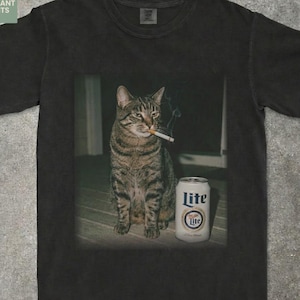 May include: Pepper-colored t-shirt featuring a cat smoking a cigarette, with a can of Lite beer beside it. The shirt has the text "FUN VIBRANT PRINTS" in the upper left corner and "PEPPER" in the lower right corner.