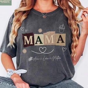 Puede incluir: Camiseta gris oscuro con la palabra "MAMA" en letras de bloque marrones, verdes y beige. La camiseta presenta un corazón, flores y un gráfico de taza de café. El texto "#Ana Lara Mathew" está debajo de la palabra "MAMA".