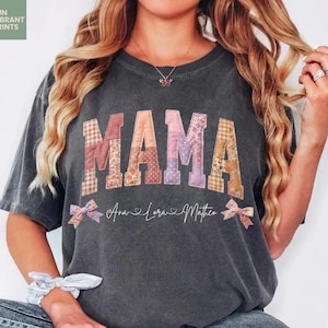 Puede incluir: Camiseta gris oscuro con la palabra "MAMA" en letras de patchwork. Las letras son de varios colores y patrones. Debajo de la palabra "MAMA" están los nombres "Ana Lora Mathew" y dos lazos rosas. La camiseta es de estilo casual.