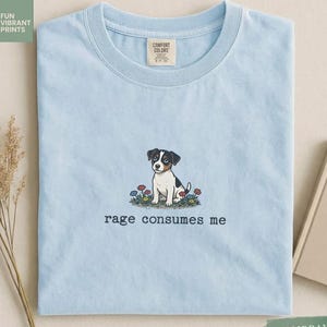 Puede incluir: Camiseta azul claro con un dibujo de un cachorro rodeado de flores y el texto "rage consumes me" debajo. La camiseta tiene cuello redondo y una etiqueta Comfort Colors. La tela es suave.
