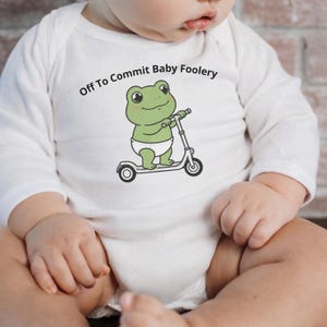 Puede incluir: Body de bebé blanco de manga larga con un dibujo de una rana verde en un patinete. La rana lleva un pañal blanco. El texto encima de la rana dice "Off To Commit Baby Foolery".