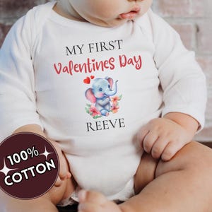 May include: White long-sleeved baby bodysuit with the text "MY FIRST Valentines Day" in black and red. A cartoon elephant with pink ears and flowers is printed on the front. The name "REEVE" is printed below the elephant. A circular graphic reads "100% COTTON".