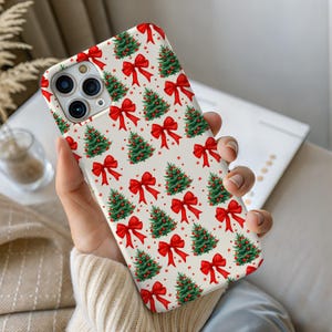 Puede incluir: Una funda de teléfono con un diseño repetido de árboles de Navidad verdes y lazos rojos sobre un fondo crema. La funda, con acabado brillante, se sostiene en la mano.