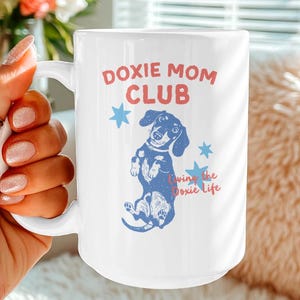 Puede incluir: Taza de cerámica blanca con el texto "Doxie Mom Club" en rojo y una ilustración de perro salchicha azul. La taza también presenta la frase "Living the Doxie Life" y gráficos de estrellas azules.