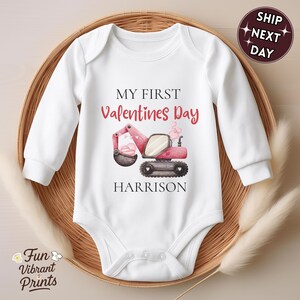 May include: White baby onesie with long sleeves, featuring a Valentine's Day design. The design includes a pink excavator with a heart-shaped bucket, the text "MY FIRST Valentines Day" and the name "HARRISON". The onesie is displayed in a woven basket.
