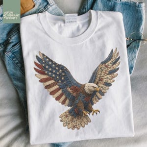 Puede incluir: Camiseta blanca con un gráfico detallado de un águila calva americana con las alas extendidas. Las alas del águila están diseñadas con las estrellas y rayas de la bandera estadounidense y patrones florales. La camiseta tiene el texto "FUN VIBRANT PRINTS" en la esquina superior izquierda.