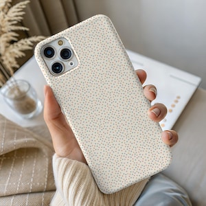 Puede incluir: Funda de teléfono de color crema con un patrón repetido de pequeños puntos multicolores. La funda se sostiene en una mano, mostrando las lentes de la cámara y el diseño general. Los puntos son en tonos azules, naranjas y beige.