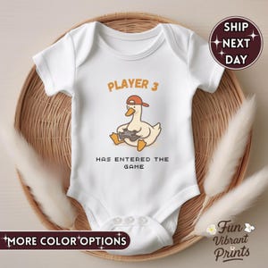 May include: White baby bodysuit with the text "PLAYER 3 HAS ENTERED THE GAME." The bodysuit features a cartoon duck wearing a red cap and holding a game controller. The bodysuit is on a woven basket. Additional text includes "MORE COLOR OPTIONS" and "Fun Vibrant Prints."