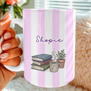 Puede incluir: Taza de cerámica blanca con un diseño de rayas rosas y blancas. La taza presenta el nombre "Shopie" en escritura púrpura, junto con ilustraciones de libros, una taza de café y una planta en maceta. La taza es sostenida por una persona.
