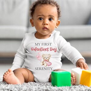 May include: A white baby onesie with long sleeves. The onesie has the text "MY FIRST Valentines Day" in red and a teddy bear holding a heart. The word "SERENITY" is below the bear. The baby is sitting on a gray carpet with colorful blocks.