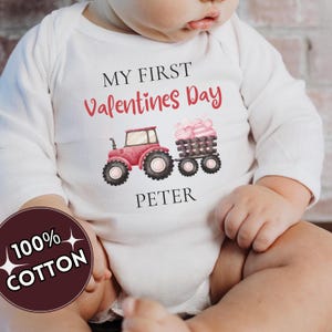 May include: White baby onesie with long sleeves. The onesie has the text "MY FIRST Valentines Day" and the name "PETER" printed on it. A red tractor with a trailer of heart-shaped candies is also printed on the onesie. The onesie is made of 100% cotton.