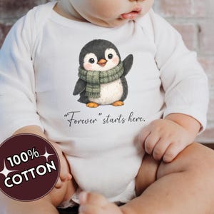 Puede incluir: Un body blanco de manga larga para bebé con el gráfico de un pequeño pingüino con bufanda verde que saluda. Debajo del pingüino, el texto dice "Forever" starts here. Una insignia circular en la esquina indica 100% COTTON.