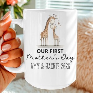 Puede incluir: Taza de cerámica blanca con una ilustración de una jirafa madre y su bebé. La taza tiene el texto "OUR FIRST Mother's Day" con los nombres "AMY & JACKIE 2026" debajo. La taza es sostenida por una persona.