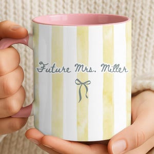 Puede incluir: Una taza de cerámica blanca con rayas verticales amarillas de acuarela y un interior rosa. La taza presenta el texto "Future Mrs. Miller" en una fuente de escritura, con una pequeña ilustración de lazo debajo.