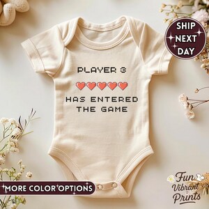 May include: A cream-coloured baby bodysuit featuring the text "PLAYER 3 HAS ENTERED THE GAME" in a black pixelated font. Above the text are five red pixelated hearts. The bodysuit has short sleeves and a round neckline. The words "MORE COLOR OPTIONS" are at the bottom.