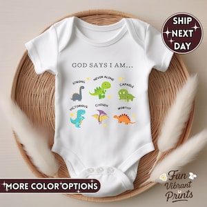 Puede incluir: Body de bebé blanco con el texto "GOD SAYS I AM..." e ilustraciones de dinosaurios coloridos. Los dinosaurios representan palabras como "Fuerte", "Nunca solo" y "Capaz". El body está sobre una cesta tejida.