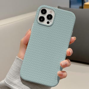 Puede incluir: Funda de teléfono azul claro con un patrón de espiga blanco repetido. La funda tiene recortes para la cámara y los botones del teléfono. La funda del teléfono está en la mano de una persona.