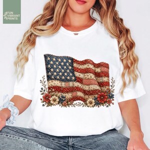 Puede incluir: Camiseta blanca con un diseño de bandera estadounidense desgastada y detalles florales. El diseño de la bandera incorpora estrellas y rayas en rojo, blanco y azul, con una estética vintage y de patchwork. La camiseta incluye el texto "FUN VIBRANT PRINTS".
