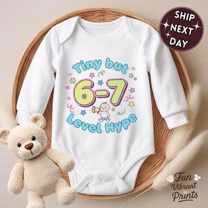 May include: White long-sleeved baby bodysuit with the text "Tiny but 6-7 Level Hype" in blue, green, and pink. The design includes stars, confetti, and a baby with a megaphone. A teddy bear and a woven basket are also in the image.