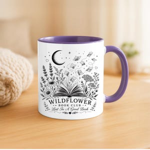 Puede incluir: Una taza de cerámica blanca con interior y asa de color morado, que presenta una ilustración en tinta negra de un libro abierto, flores silvestres, una luna creciente y páginas flotantes. El texto dice: WILDFLOWER BOOK CLUB LOST IN A GOOD BOOK.