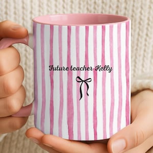 Puede incluir: Una taza de cerámica a rayas rosas y blancas con interior y asa rosas. La taza presenta el texto "Future teacher Kelly" y un lazo negro. La taza está sujeta por una persona.