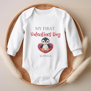 May include: White baby onesie with long sleeves, featuring the text "MY FIRST Valentines Day" in black and red. A cartoon penguin is centered on a red heart, with the name "SOPHIA" below. The onesie is displayed in a woven basket.