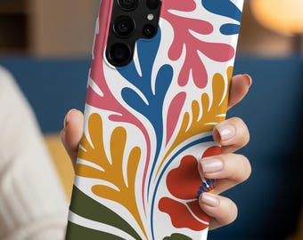 Pode incluir: Uma capa de telefone branca com um design floral colorido. O padrão inclui formas de folhas vermelhas, rosa, azuis, amarelas e verdes. A capa do telefone está em uma mão.