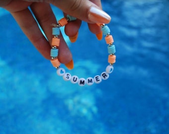 May include: A beaded bracelet with the word "SUMMER" spelled out in white beads with a blue heart on each end. The bracelet is made with alternating blue and orange beads.