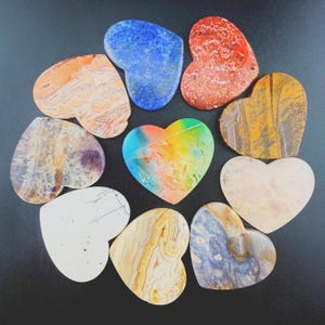 May include: A collection of ten heart-shaped stones in various colors and patterns, including blue, red, orange, yellow, white, and purple, arranged in a circular formation. Each stone features unique natural textures, mineral veins, and color gradients.