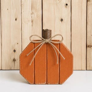 May include: A flat, orange, octagonal wooden pumpkin decoration featuring vertical slats and a natural brown twig stem. A thin, light brown jute twine is tied into a bow around the stem, resting against the front of the orange wooden surface.