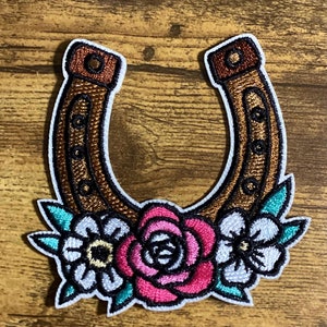 May include: A brown horseshoe with a white outline and gold accents is decorated with three embroidered flowers. The flowers are pink, white, and teal.