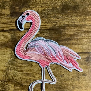 May include: Pink flamingo embroidered iron-on patch with white outline. The flamingo is standing on one leg with its head bent down.