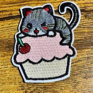 May include: Embroidered iron-on patch featuring a gray cat with red cheeks sitting on top of a pink cupcake with a red cherry on top.
