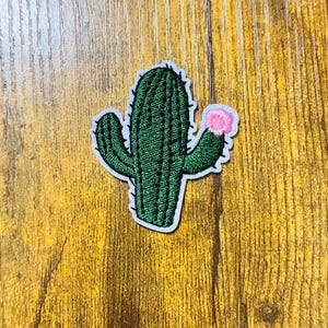 May include: Embroidered cactus patch with a green body, black details, and a pink flower. The patch has a white border and is set against a wooden background. This patch is ideal for adding a desert-themed touch to clothing or accessories.