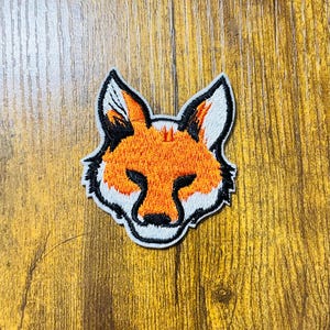 May include: Embroidered iron-on patch of a fox's face. The fox has orange fur, black eyes, and a white muzzle.