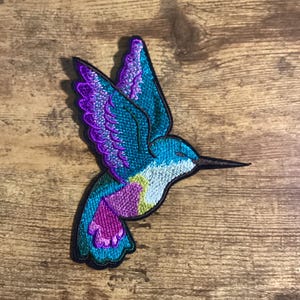May include: A colorful embroidered hummingbird patch with purple, blue, and green accents. The patch is perfect for adding a touch of nature to clothing, bags, or other accessories.