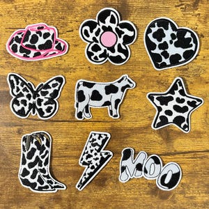 May include: A collection of embroidered patches with a cow print design. The patches include a cowboy hat, flower, heart, butterfly, cow, star, cowboy boot, lightning bolt, and the word "MOO". The patches have a white border and are on a wooden surface.
