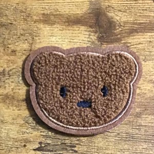 May include: Brown fuzzy bear face patch with a stitched black nose and eyes. The patch is outlined in a lighter brown thread.