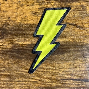May include: A neon yellow lightning bolt patch with a black border. The patch is made of fabric and has a textured appearance. The lightning bolt design is a classic symbol of energy and speed.