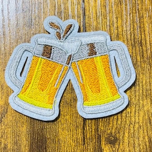 May include: Two embroidered beer mugs clinking together, a fun and festive design for a patch or iron-on.