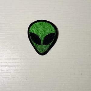 May include: Embroidered patch featuring a green alien head with black eyes and a black border. The patch has a textured appearance and is likely designed to be ironed or sewn onto fabric. The alien design is a popular theme.