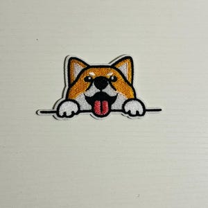 May include: Embroidered patch featuring a Shiba Inu dog with orange and white fur, black nose, and a red tongue sticking out. The dog is peeking over a black line, with white paws visible.