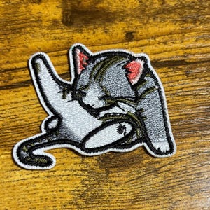 May include: Embroidered iron-on patch featuring a gray cat curled up in a sleeping position. The cat has pink ears and a black outline.