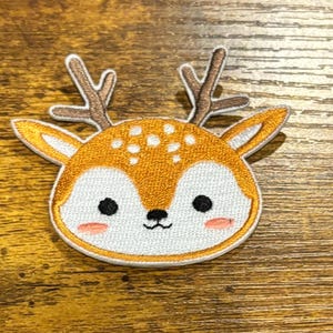 May include: Embroidered patch featuring a cartoon deer head. The deer has brown antlers, a tan and white face, and black eyes. The patch is outlined in white and orange, with a light brown background.