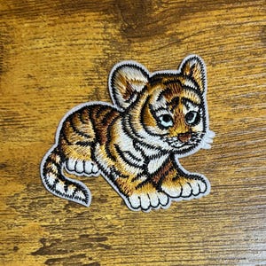 May include: A small, embroidered patch of a cute tiger cub with brown and white stripes. The tiger cub is sitting on its hind legs with its front paws raised. The patch is on a wooden background.